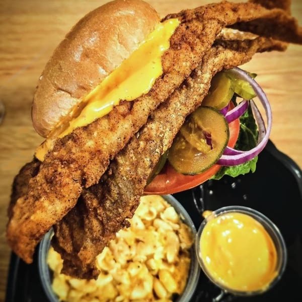 A fried fish sandwich with cheese, pickles, onions, and lettuce, served with mac and cheese and sauce.
